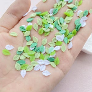 May include: A collection of small, leaf-shaped embellishments in shades of green and white. The leaves vary in color from light to dark green, with some in a bright white. These decorative elements are likely used for crafts or nail art.