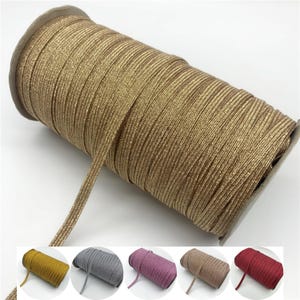 May include: A spool of gold elastic cord. The cord is tightly wound and has a slight sheen. Several smaller spools of elastic cord in yellow, grey, pink, tan, and red are shown below the main spool.