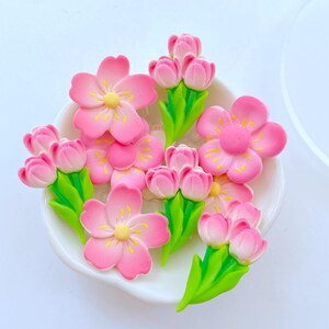 May include: A collection of pink flower and tulip-shaped embellishments with yellow centers and green stems. The flowers are arranged in a white, shallow dish, creating a decorative floral display. The image is brightly lit.