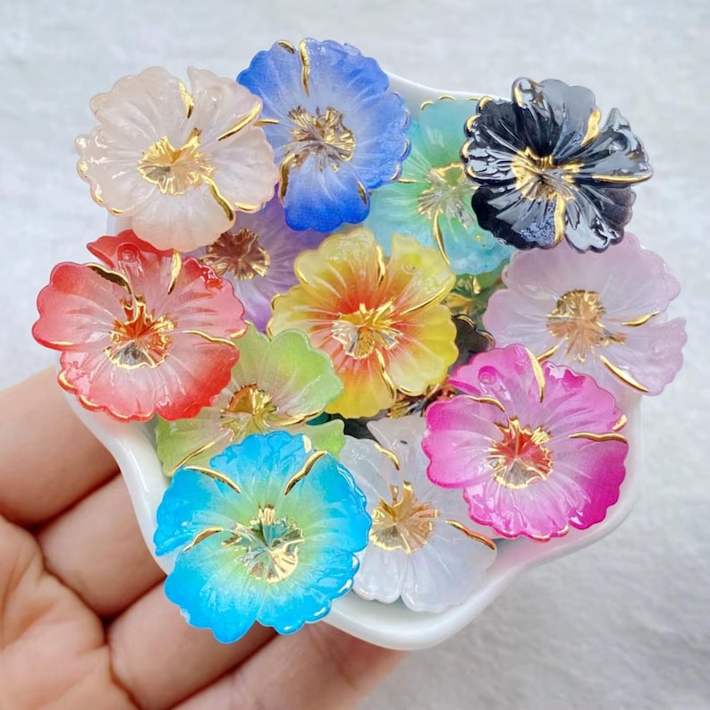 May include: A collection of colorful, translucent flower-shaped beads. Each bead features a different color, including white, blue, pink, and yellow, with gold-colored accents. The beads are arranged in a white, scalloped dish.