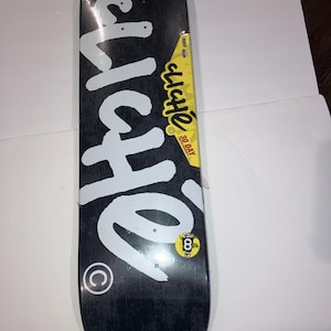 May include: A black skateboard deck featuring the white text Cliche in a large script font. A yellow sticker on the side displays the text 30 DAY LIMITED WARRANTY, and a small circular sticker near the bottom shows the number 8.