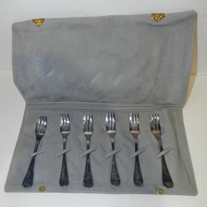May include: A set of six silver forks displayed in a gray, suede-like case. The case is open, revealing the forks secured by gray straps. The case has a gold clasp and button closure.