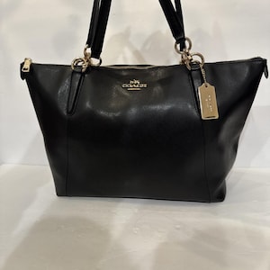 May include: A black leather tote bag featuring gold-tone hardware, two shoulder straps, and a top zipper closure. The front displays a gold Coach logo and a matching rectangular leather hangtag attached to a gold ring on the right side.