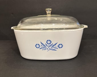 May include: A white oval casserole dish with a clear glass lid. The exterior of the white dish features a blue floral pattern consisting of three stylized flowers with leaves centered on the front side. The dish has small handles on each side.
