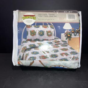 May include: A 3-piece twin sheet set made of 100% cotton flannel featuring a Teenage Mutant Ninja Turtles print. The packaging displays a white bed frame with a pillowcase and bed sheet decorated with circular character graphics on a white background.