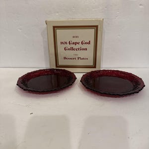 May include: Two round, deep red glass dessert plates with a textured, scalloped rim pattern, accompanied by a square cardboard box labeled Avon 1876 Cape Cod Collection Two Dessert Plates, featuring a gold and red border design on the packaging.