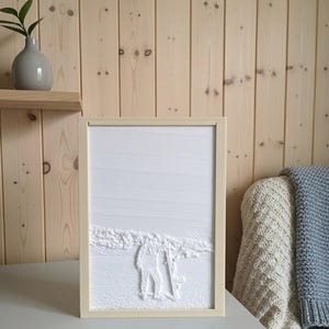 Puede incluir: Escultura en relieve blanca enmarcada que representa a una persona esquiando. La obra de arte está enmarcada en un marco de madera de color beige claro. El fondo es una superficie blanca texturizada, sobre una mesa gris.