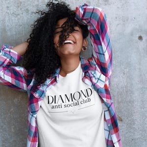 May include: A white t-shirt with the text "DIAMOND anti social club" in black. The person is wearing a plaid shirt with red, blue, and white colors. The background is a gray concrete wall.
