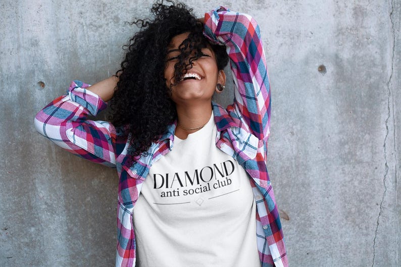 May include: A white t-shirt with the text "DIAMOND anti social club" in black. The person is wearing a plaid shirt with red, blue, and white colors. The background is a gray concrete wall.