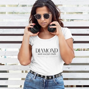 May include: A white t-shirt with the text "DIAMOND anti social club" in black. The person in the image is wearing sunglasses and holding black headphones. The person is wearing blue jeans and a black belt.