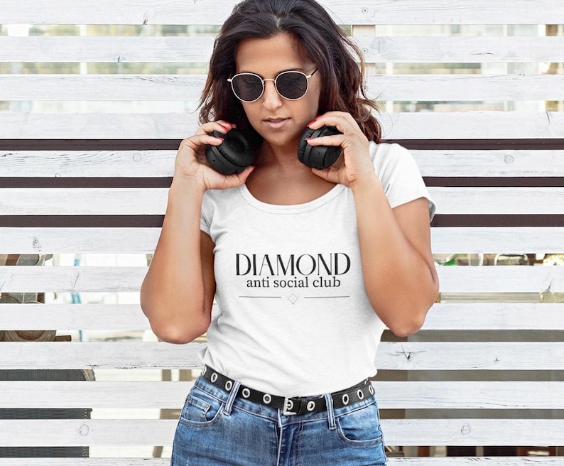 May include: A white t-shirt with the text "DIAMOND anti social club" in black. The person in the image is wearing sunglasses and holding black headphones. The person is wearing blue jeans and a black belt.