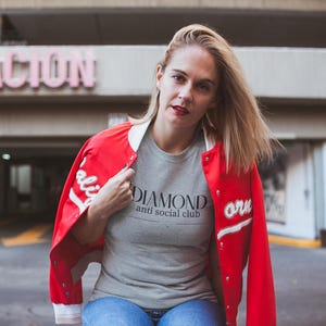May include: A woman wearing a red and white letterman jacket over a gray t-shirt and blue jeans. The t-shirt has the text "DIAMOND anti social club". The jacket has white lettering on the sleeve. The background includes a building with the word "STACION" in pink letters.