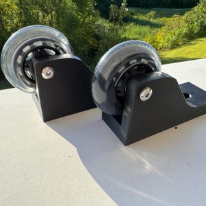 May include: Two black roller skate wheel assemblies with clear wheels. The wheels have a black inner core and are mounted on black brackets with silver hardware. The wheels are on a white surface with a green background.