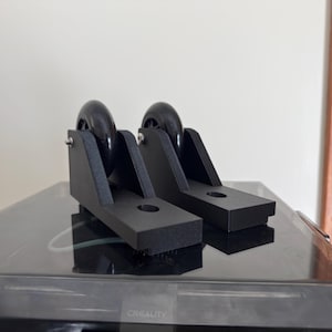May include: Two black 3D-printed caster wheel assemblies. Each assembly features a black wheel, mounted on a black bracket with a mounting hole. The assemblies are designed for use on a 3D printer or similar equipment.