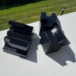 May include: Three black, solid, block-shaped stands with a curved, concave indentation on the top surface. Each stand features a stepped, tiered design with smooth, glossy surfaces and sharp, angular edges, arranged in a cluster on a flat surface.