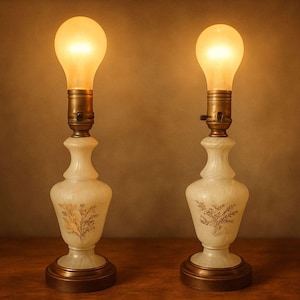 Może przedstawiać: Dwie zabytkowe lampy stołowe z podświetlonymi żarówkami. Lampy mają kremową, ozdobną podstawę ze złotymi akcentami i mosiężną szyjką. Żarówki mają ciepły żółty kolor, rzucając miękką poświatę. Lampy stoją na drewnianej powierzchni.