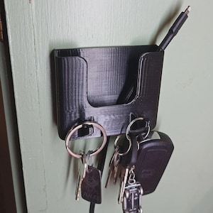 May include: A black 3D-printed key holder mounted on a wall. The holder has a slot for keys and a separate compartment for a charging cable. Several keys and a car key fob are hanging from the holder.