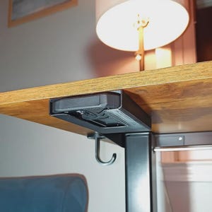 May include: A black under-desk phone holder and hook attached to a wooden desk. The phone holder is rectangular and mounted to the underside of the desk. A curved hook hangs below the holder. A lamp and chair are in the background.