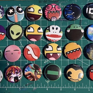May include: A collection of 24 round button pins with various colorful designs. The pins feature cartoon characters, logos, and abstract art. The pins are arranged on a green cutting mat.