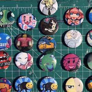 May include: A collection of twenty-one colorful, round pinback buttons with various cartoon character designs. The buttons feature a variety of colors, including blue, green, red, and pink. Some buttons have text, and the buttons are arranged on a green cutting mat.