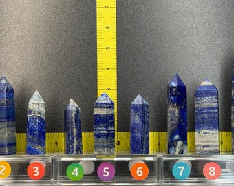 May include: Nine lapis lazuli crystal towers arranged in a row, numbered one through nine. A yellow measuring tape is visible behind the stones, showing heights ranging from approximately 2.5 to 5.5 inches. Each stone features blue and white patterns.