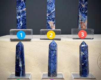 May include: Six polished sodalite crystal towers arranged in two rows of three. Each stone features deep blue hues with white and occasional orange-brown mineral inclusions. Each tower is mounted on a clear rectangular base labeled with a numbered circle.