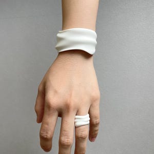 Puede incluir: Un conjunto de pulsera y anillo blancos texturizados. La pulsera es ancha y se envuelve alrededor de la muñeca. El anillo está en un dedo. Ambas piezas tienen un diseño ondulado y orgánico. La joyería está hecha de un material opaco.