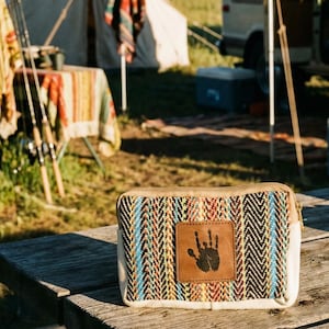 Puede incluir: Un estuche rectangular con cremallera y un patrón de espiga tejido en azul, negro, rojo y beige. Un parche de cuero marrón con una huella de mano negra está centrado en la parte delantera. El estuche está sobre una superficie de madera.