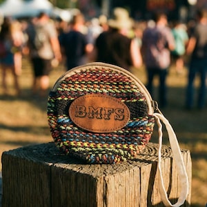 Puede incluir: Un monedero redondo tejido con un parche de cuero marrón que muestra las letras "BMFS". El monedero tiene un cierre de cremallera y una correa de muñeca color crema. El patrón tejido incluye hilos negros, rojos, amarillos y azules.