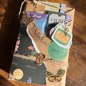 May include: A wrapped book package featuring a black floral paper overlay with the word Thriller written in script. It includes a moth sticker, a wax seal, a Yogi tea bag, a reading list card, and a sticker that reads Smells like she's reading thrillers again.