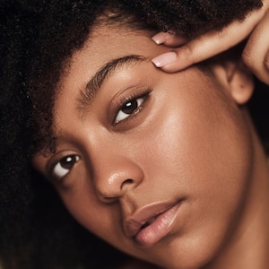 May include: Close-up portrait of a person with dark skin and curly black hair. The person is looking at the camera with a neutral expression. A finger is touching the eyebrow. The skin has a healthy glow.