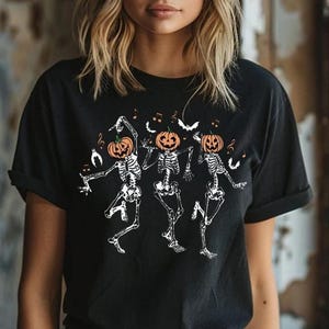 May include: A black short-sleeve t-shirt featuring a graphic print of three white skeletons with orange pumpkin heads dancing. The design includes small white bats and musical notes scattered around the skeletons on the front of the shirt.