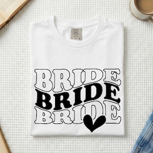 May include: A white short-sleeved t-shirt featuring a black graphic print with the word BRIDE repeated three times in a wavy, stacked font. The middle word is solid black, while the top and bottom words are outlined. A solid black heart is below.
