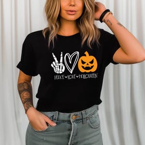 May include: A black short-sleeved t-shirt featuring a white graphic of a skeleton hand making a peace sign, a white heart outline, and an orange jack-o'-lantern pumpkin. Below the graphics, the text reads PEACE LOVE HALLOWEEN in white.