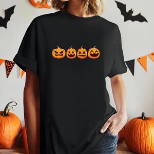 Może przedstawiać: Czarny t-shirt z krótkim rękawem z nadrukiem graficznym przedstawiającym cztery pomarańczowe dynie w poziomym rzędzie. Każda dynia ma inny wyrzeźbiony wyraz twarzy, w tym złą minę, klasyczną dynię na Halloween, twarz z oczami w kształcie X oraz szeroki uśmiech.