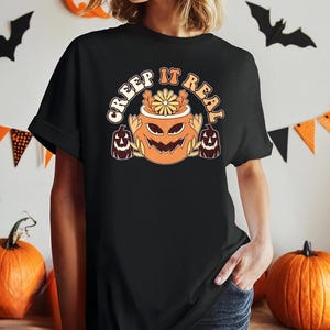 May include: A black short-sleeved t-shirt featuring a graphic print of a central orange jack-o'-lantern with a flower on top, flanked by two smaller dark brown jack-o'-lanterns, with the text CREEP IT REAL arched above in a light cream color.