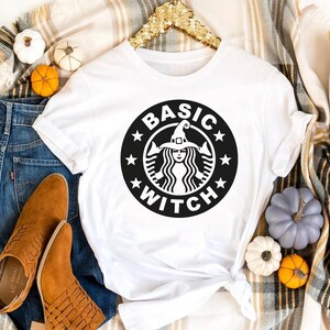 May include: A white short-sleeved t-shirt featuring a black circular graphic on the chest. The graphic depicts a stylised figure wearing a witch hat, surrounded by the text "BASIC WITCH" and small stars, mimicking a coffee shop logo design.