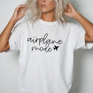 May include: A white short-sleeved t-shirt featuring the black text "airplane mode" written in a cursive script font, accompanied by a small black silhouette of an airplane icon positioned to the right of the word mode on the chest area.