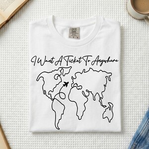 May include: A white t-shirt featuring a black line-art world map graphic. Above the map, the text reads I Want A Ticket To Anywhere in a cursive font. A small black airplane icon is positioned over the Atlantic Ocean with a dotted flight path.