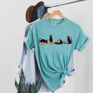 May include: A teal short-sleeve t-shirt featuring a graphic print of four black cats interacting with orange pumpkins and a skull. The shirt is displayed on a wooden hanger, layered over a light blue denim shirt and a brown hat.
