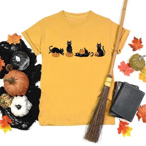 Peut inclure: Un t-shirt à manches courtes jaune moutarde avec une impression graphique représentant quatre chats noirs interagissant avec des citrouilles orange et un crâne. Les chats sont représentés dans diverses poses, assis, couchés ou jouant avec les courges.
