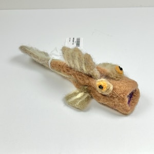 May include: A needle-felted fish figurine in shades of tan and beige with prominent textured fins, a long tail, and two large, round yellow eyes with black pupils. The fish has an open mouth revealing a purple interior and a small white product tag.