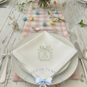 May include: A beautifully set Easter table with a white monogrammed napkin, a pink and white checkered runner, and a vase of flowers. Decorative Easter eggs and a bunny figurine add to the festive decor. Silverware and crystal glasses complete the elegant setting.