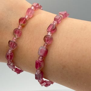 May include: Two pink tourmaline bracelets. The bracelets are made of small, irregular, faceted beads and round beads strung together. The color of the beads is a deep pink with some lighter pink and clear inclusions. The bracelets are displayed on a person's arm.