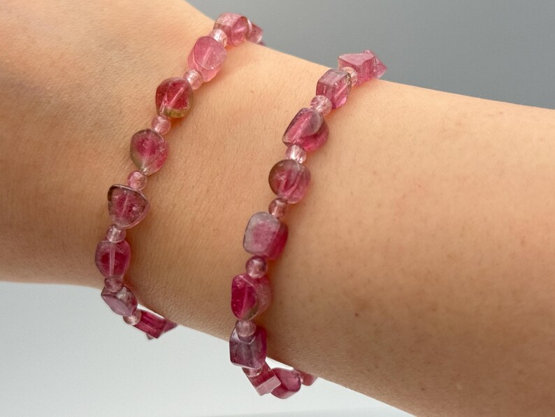 May include: Two pink tourmaline bracelets. The bracelets are made of small, irregular, faceted beads and round beads strung together. The color of the beads is a deep pink with some lighter pink and clear inclusions. The bracelets are displayed on a person's arm.