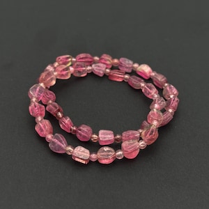 May include: A double-strand bracelet featuring pink tourmaline beads. The beads are irregular in shape and vary in shades of pink, from light to deep rose. The bracelet is displayed against a dark background.