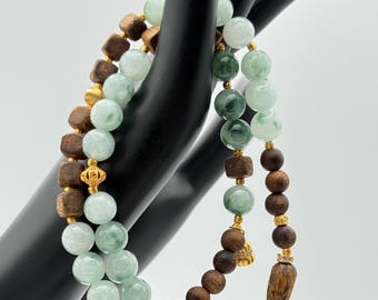 May include: Two beaded necklaces. One features light green jade beads and wooden cube beads with gold accents. The other has dark brown wooden beads and a wooden pendant with gold accents. The necklaces are displayed on a black hand.