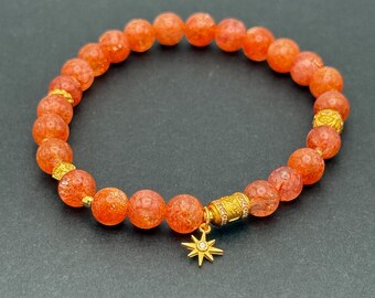 May include: Orange beaded bracelet with gold accents and a star charm. The bracelet features round, textured beads and gold-colored spacers. A gold-colored charm in the shape of a star hangs from the bracelet. The bracelet is displayed on a dark gray background.