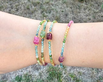 May include: A collection of beaded bracelets in various colors, including pink, green, and purple. The bracelets are strung with small, round beads and feature larger, irregularly shaped gemstone accents. The bracelets are worn on a wrist.