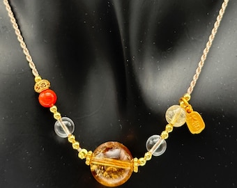 May include: A gemstone necklace featuring a large, round, amber-colored bead as the focal point. The necklace includes smaller clear and orange beads, gold accents, and a gold tag. The cord is a light beige color.